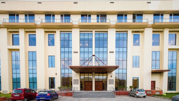 A modern building with a large entrance, multiple windows, and a parking area with several cars. - Eternal Oasis by Alaya Stays (Gurugram)