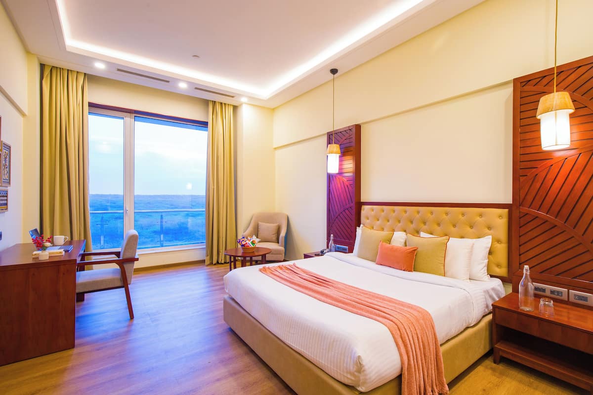 A hotel room with a large bed, a desk, a chair, a small table, and a nightstand. There is a window with curtains and a view of the city.