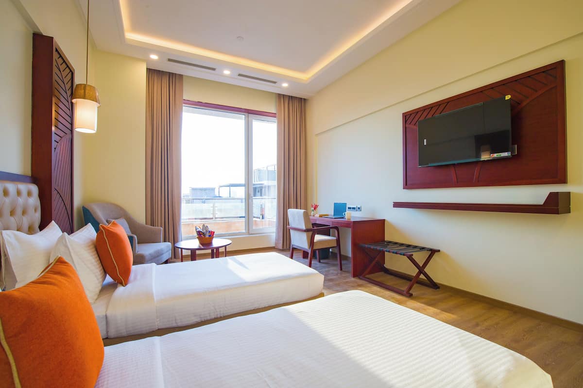 A hotel room with two beds, a desk, a chair, a TV mounted on the wall, and a view of the city through large windows.