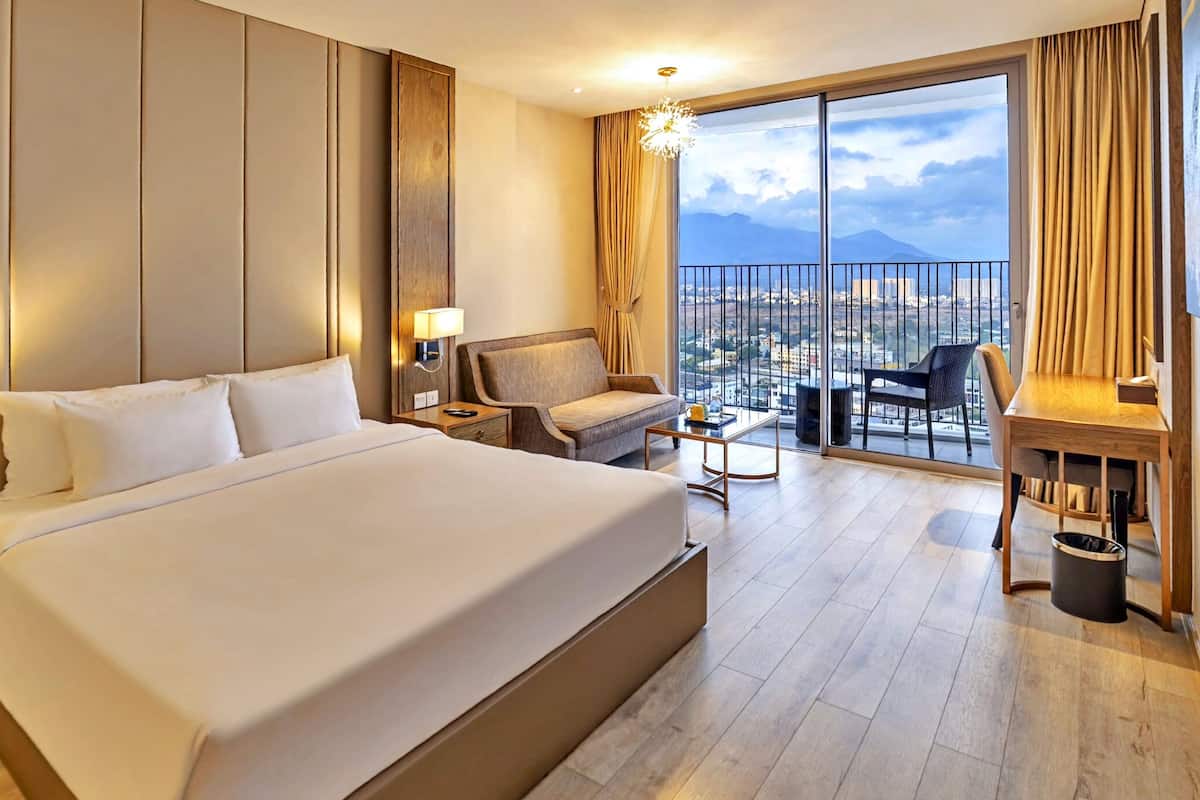 A hotel room with a large bed, a desk, a chair, and a balcony with a city view.