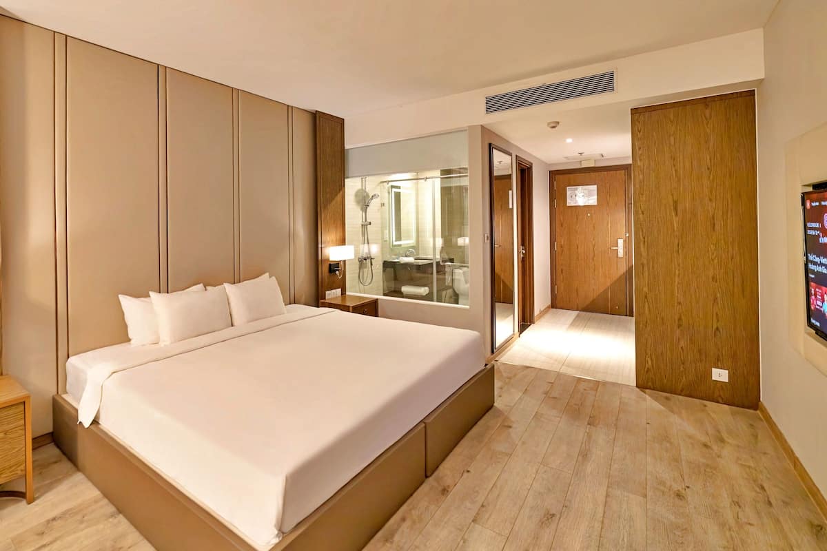 A modern hotel room with a large bed, wooden paneling, a nightstand with a lamp, a mirror, and a door leading to a bathroom.
