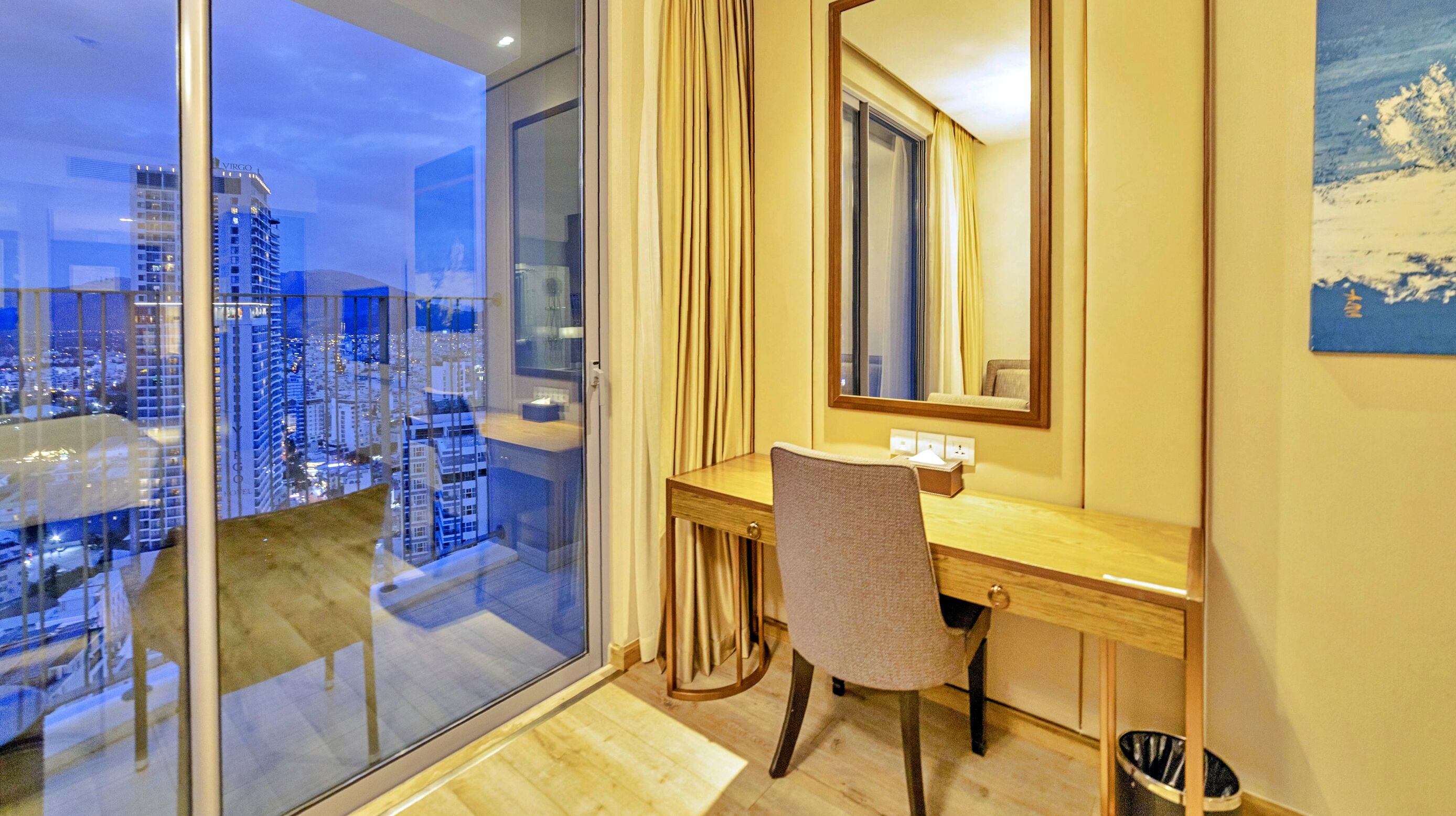 A hotel room with a large window, a wooden desk, a chair, a mirror, and a view of the cityscape.