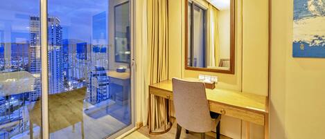 A hotel room with a large window, a wooden desk, a chair, a mirror, and a view of the cityscape.