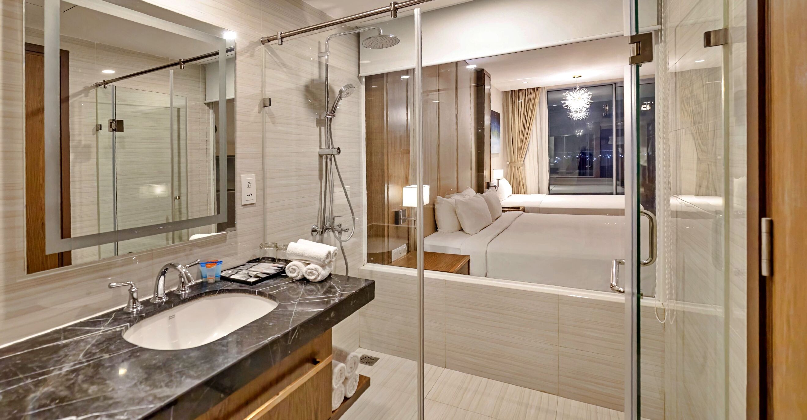 A modern bathroom with a glass-enclosed shower, a large mirror, a marble sink, and a bedside table with a lamp.