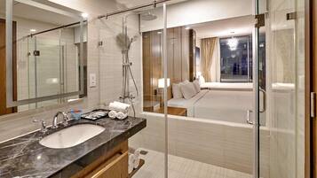 A modern bathroom with a glass-enclosed shower, a large mirror, a marble sink, and a bedside table with a lamp.