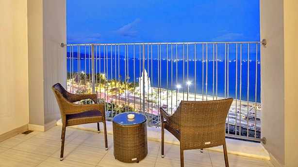 Een balkon met twee rieten stoelen en een tafeltje, met uitzicht op een stadsgezicht en de zee en verre gebouwen.
