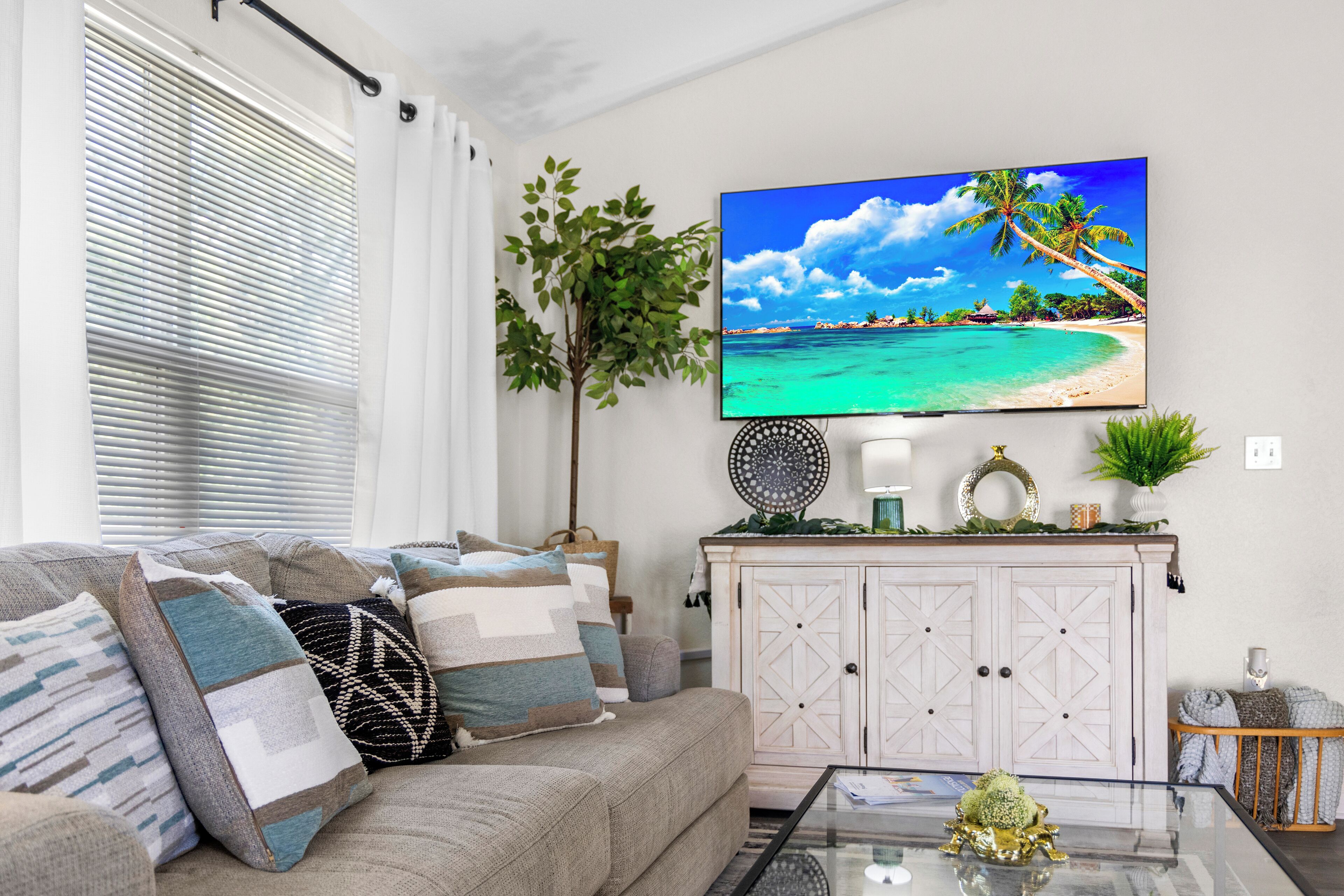 Uma sala de estar com uma televisão de tela plana grande exibindo uma cena de praia, um sofá com almofadas estampadas, uma mesa de centro de vidro com um objeto decorativo e um armário branco com objetos decorativos.