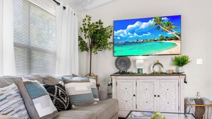 Una sala de estar con un televisor de pantalla plana grande mostrando una escena de playa, un sofá con cojines estampados, una mesa de centro de vidrio con un objeto decorativo y un armario blanco con elementos decorativos.