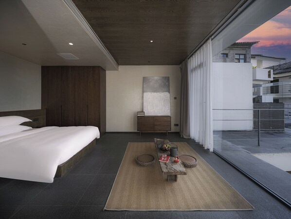 Un dormitorio moderno con una cama grande, un armario de madera, una mesita de noche con lámpara y vistas a la ciudad a través de amplios ventanales.