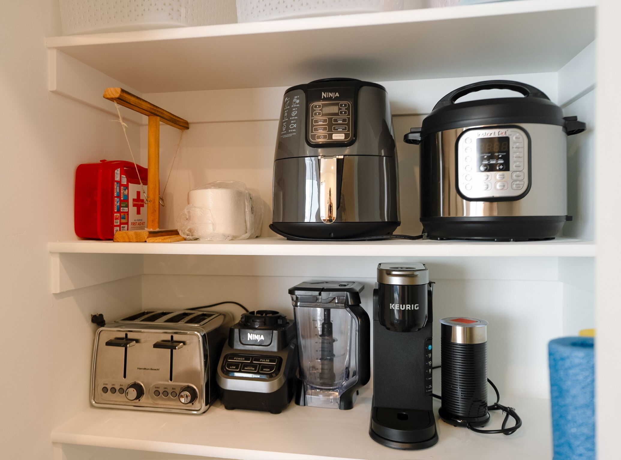 Une étagère de cuisine avec un grille-pain, un mixeur de la marque Ninja, une machine à café Keurig, un cuiseur à riz et une trousse de premiers secours rouge.