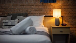 A neatly made bed with pillows and a rolled towel, a bedside lamp, a nightstand with a clock, and a brick wall background. - Queen Deluxe Retreat, Smart TV, Kitchenette & More! (Coffs Harbour)