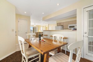 Un comedor con una mesa de madera y sillas blancas, una cocina con armarios y un microondas, y una puerta con ventana.