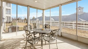 A covered patio with a round table and chairs, large windows, and a view of a white fence and buildings.