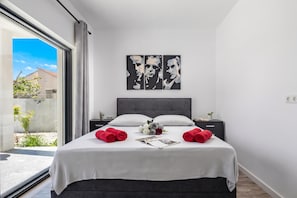 A modern bedroom with a large bed, two nightstands, a grey headboard, and a window with a view of a building and greenery. - Villa Claudio With Private Pool (Vodice)