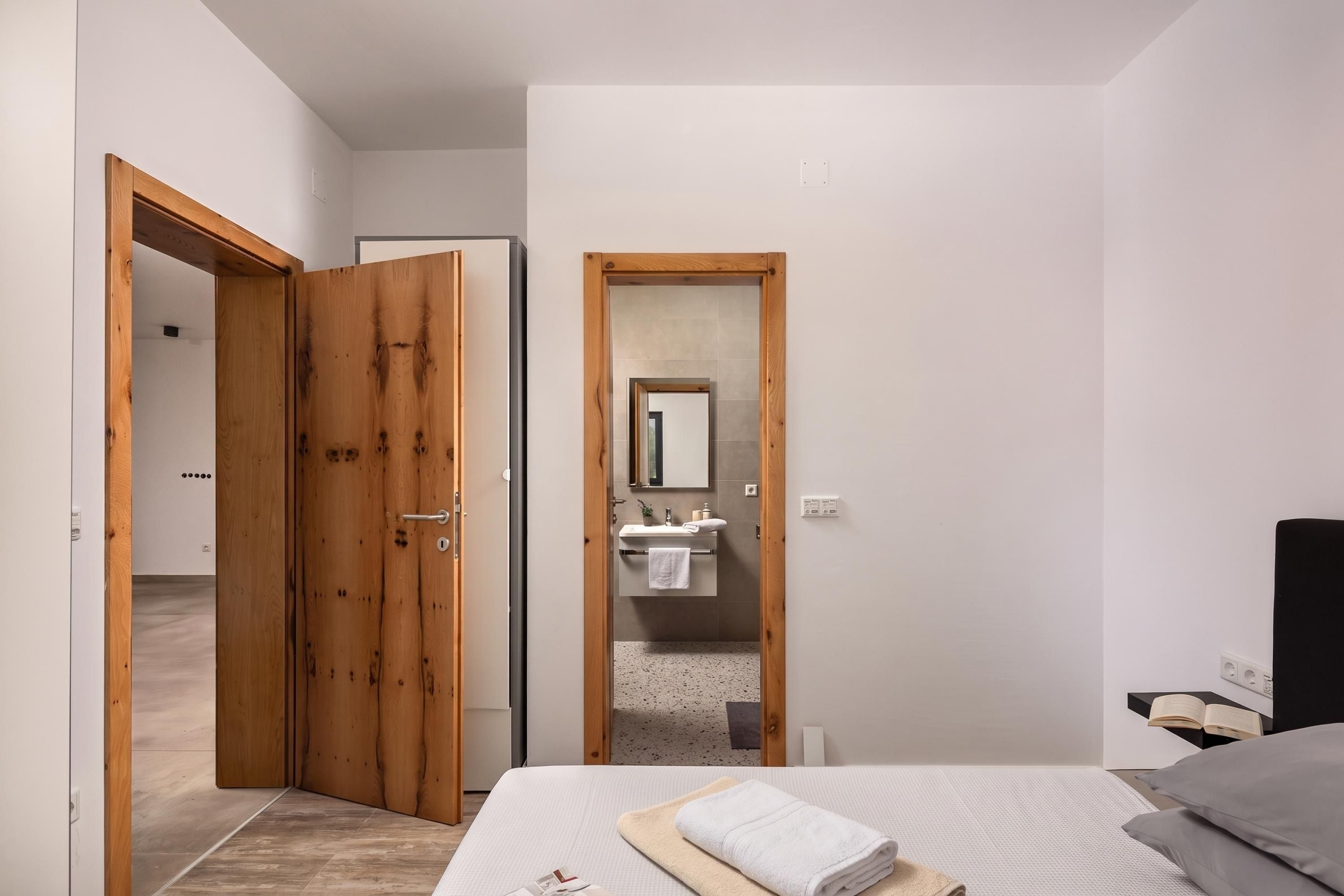 A bedroom with a bed, a bedside table with a book, a wooden door, and a bathroom visible through a mirror.