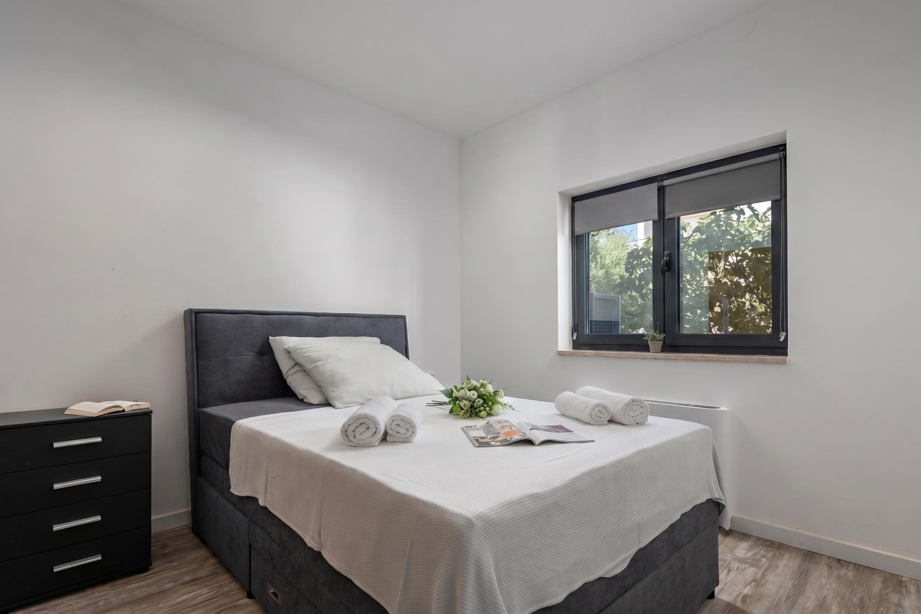 A modern bedroom with a bed, a nightstand, a window, and a view of trees.