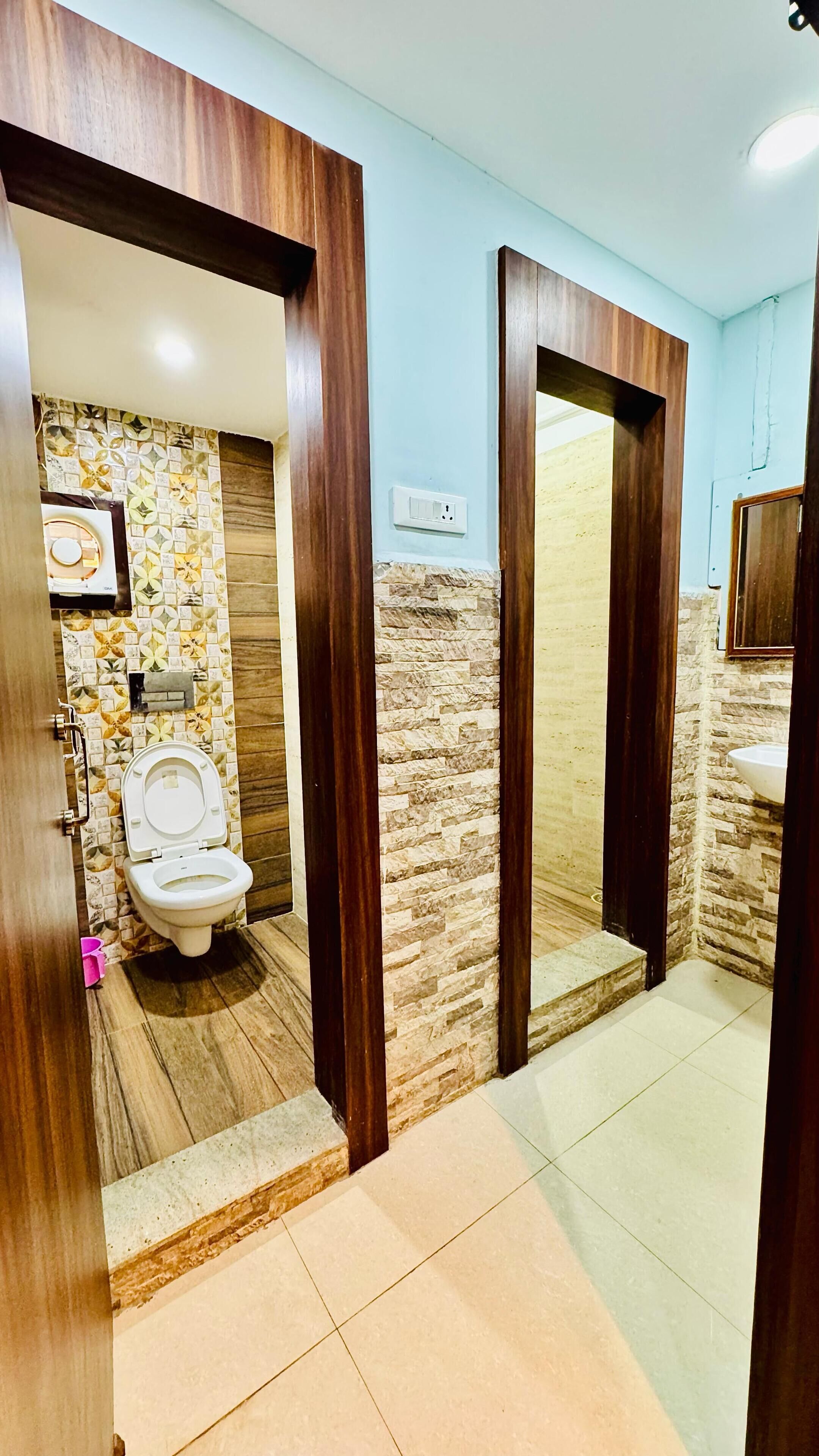 Un baño con inodoro, lavamanos y espejo. Se aprecia un marco de puerta de madera y suelo de baldosas.