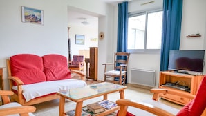 Ein gemütliches Wohnzimmer mit einem roten Sofa, Holzstühlen, einem Glastisch mit Büchern, einem Fernseher auf einem Holzboden und einem großen Fenster mit blauen Vorhängen.