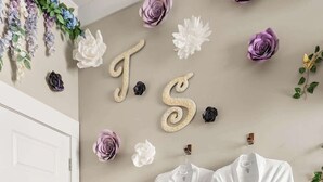 Une pièce avec un plancher en bois, une porte blanche et un mur décoré de fleurs et de lettres en plastique. Deux robes blanches sont accrochées au mur.