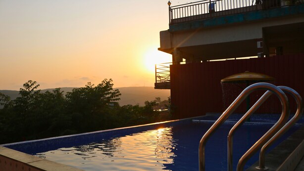 A swimming pool with a metal railing, a view of the sunset, and a building in the background. - Melody 24 East Resort (Mahabaleshwar)