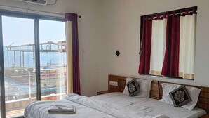 A hotel room with a large bed, a ceiling fan, a window with curtains, and a view of a pool and building. - Melody 24 East Resort (Mahabaleshwar)