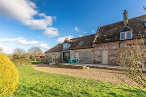 Une maison en pierre avec un toit en pente, un porche à l’avant orné de chaises vertes et une pelouse bien entretenue.