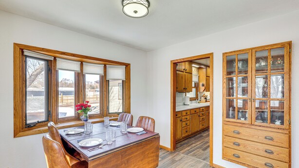 Un coin repas avec une table et des chaises en bois, un vase rempli de fleurs, et une cuisine dotée de meubles en bois et d’un placard intégré avec des portes en verre.