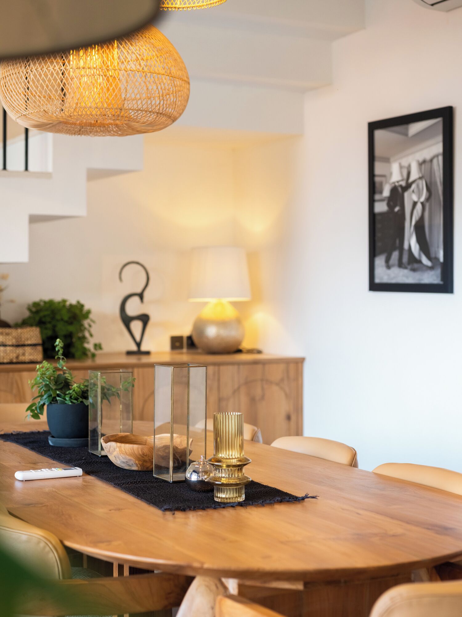 A dining area with a wooden table, clear glass candle holders, a potted plant, a lamp, and a framed picture on the wall.