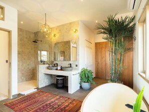 Une salle de bain moderne avec un grand miroir, deux lavabos, une douche et une baignoire. On y trouve également une plante en pot et un tapis sur le sol.