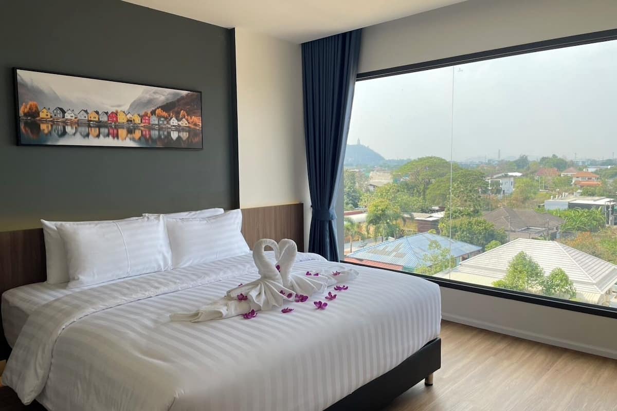 A hotel room with a large bed, a painting on the wall, and a view of the city through a large window.