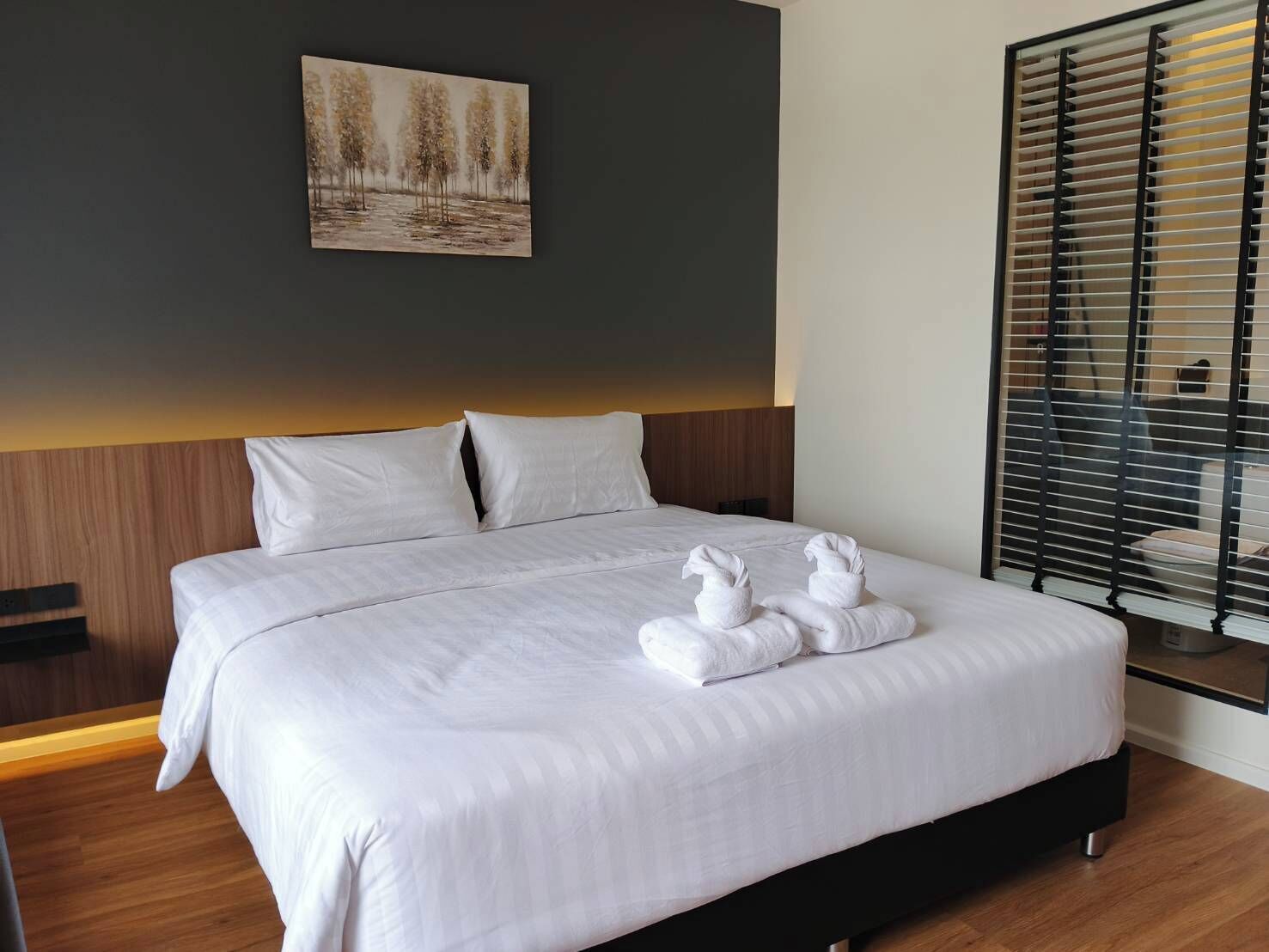 A neatly made bed with white linens and two towels folded on top, a painting of trees on the wall, and a window with blinds.