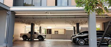 Un estacionamiento techado con dos coches aparcados, un edificio con balcón y un árbol.