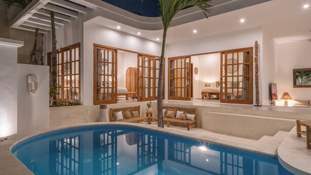 A modern pool area with a clear blue pool, wooden furniture, and large windows with wooden frames.