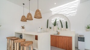 A modern kitchen with a central island, wooden stools, hanging wicker pendant lights, and potted plants.