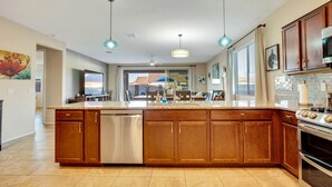A spacious kitchen with wooden cabinets, a stainless steel dishwasher, and a large island with a sink. There are pendant lights above the island and a dining area in the background.