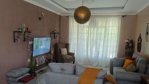 Uma sala de estar com um sofá cinzento, uma poltrona estampada, uma televisão e uma luminária pendente decorativa.