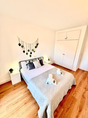 Ein ordentlich bezogenes Bett mit grauer Decke, ein Nachttisch mit Lampe und eine Wanddekoration.