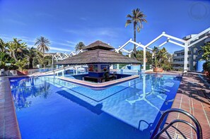 Ein Swimmingpool mit einer strohgedeckten Struktur, Palmen und blauem Himmel.