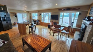 Amplia sala de estar con una mesa de centro de madera, una cocina con refrigerador, y ventanas grandes que ofrecen vistas al exterior.