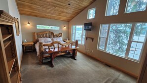 Un dormitorio con cama de madera, televisor montado en la pared y ventanas amplias con vistas a árboles.