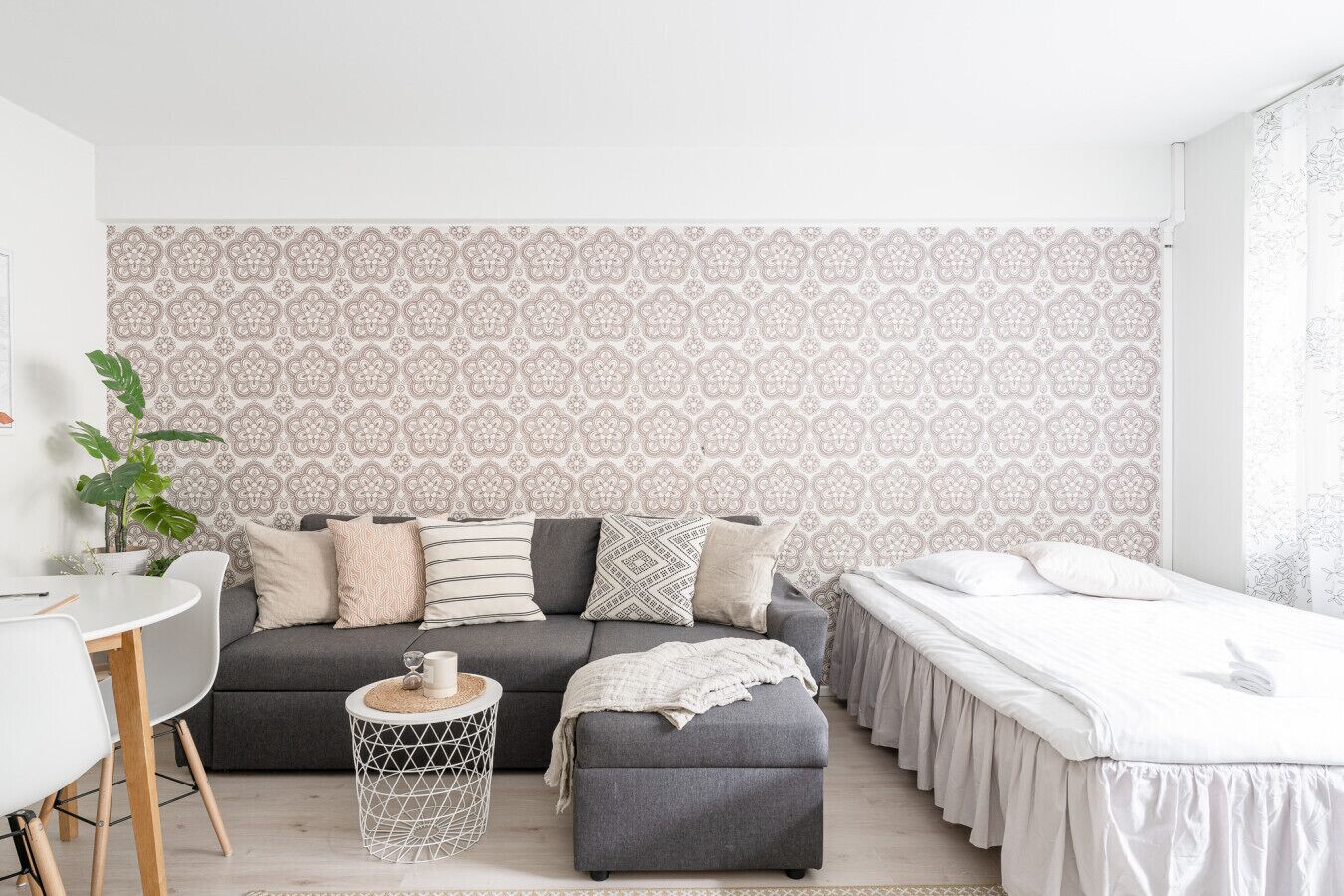 A modern bedroom with a grey sofa, a small ottoman, a bed with white bedding, and a patterned wallpaper.