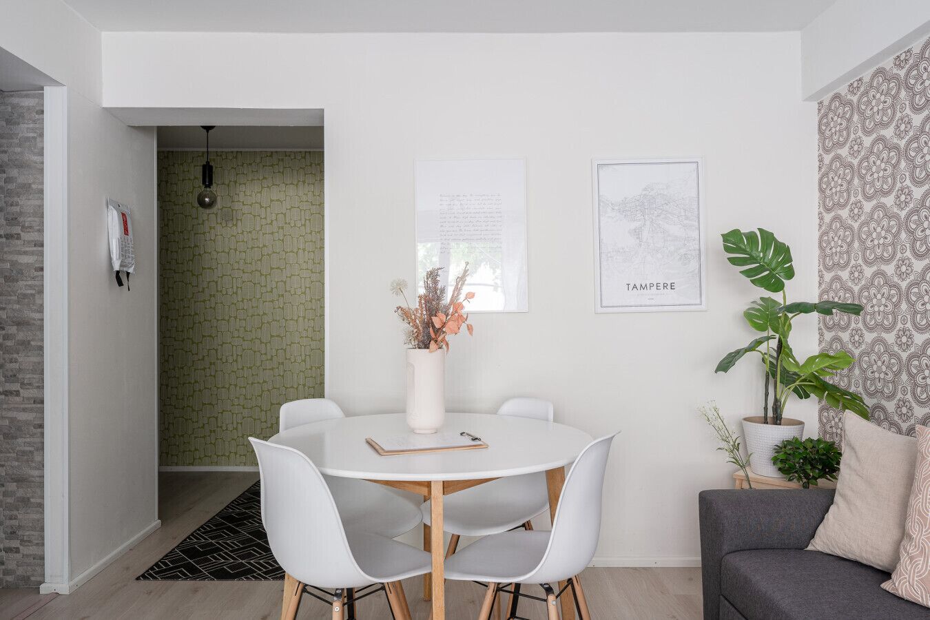 Un coin repas moderne avec une table ronde, des chaises blanches et un vase contenant des fleurs séchées. Deux tableaux encadrés sont accrochés au mur, dont l’un porte l’inscription « TAMPERE ».