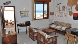 A living room with a sofa, two wooden chairs, a coffee table, a small table with a lamp, and a view of the beach through a large window. - ON THE WATERFRONT UNIQUE LOCATION!!!! (San Vito Lo Capo)