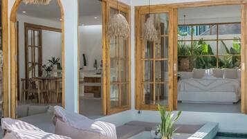 Un área de piscina moderna al aire libre con zona de estar, mesa de comedor y un dormitorio visible a través de amplios ventanales.