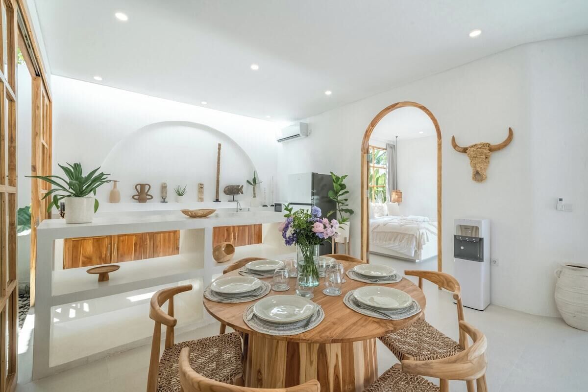 Un moderno comedor con una mesa redonda de madera dispuesta para cuatro personas, rodeada de sillas de mimbre. Se ve una cocina blanca con mostrador de madera al fondo.