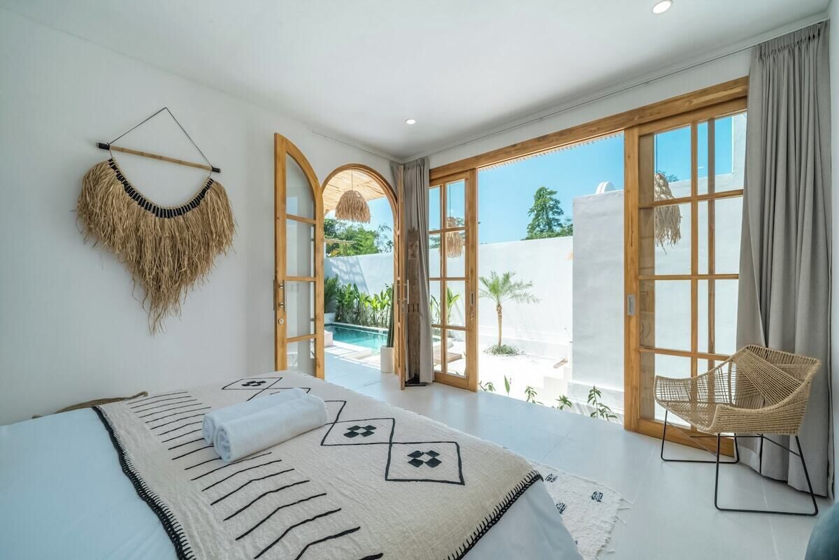 A spacious bedroom with a large bed, a wicker chair, and a view of a pool and greenery.