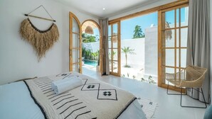 Ein geräumiges Schlafzimmer mit einem großen Bett, einem Korbstuhl und Blick auf einen Pool und Grünflächen.