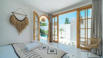 A spacious bedroom with a large bed, a wicker chair, and a view of a pool and greenery.
