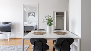 Une pièce moderne et minimaliste, avec une table à manger blanche, deux chaises noires, un petit vase blanc contenant des plantes vertes, un canapé gris, un lit avec une literie blanche et un téléviseur posé sur un meuble blanc.