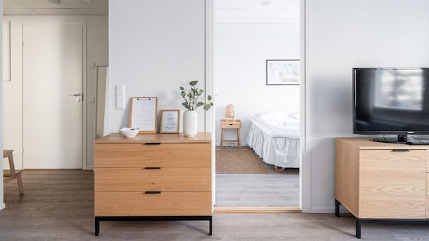 A modern hotel room with a wooden dresser, a television on a wooden stand, a bed with white bedding, and a small bedside table with a lamp.